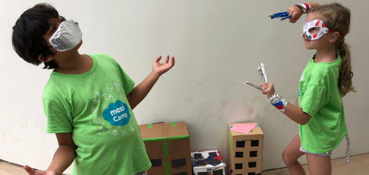 two children in green MOXI camp shirts pose like superheroes with cardboard masks