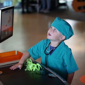 young boy dressed in scrubs with a stethoscope around his neck looks up at the MOXI globe with his hand on the sensor