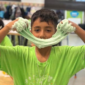 young boy wearing a bright green shirt stretches green slime in front of his face like a smile