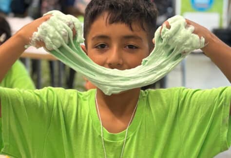 young boy wearing a bright green shirt stretches green slime in front of his face like a smile