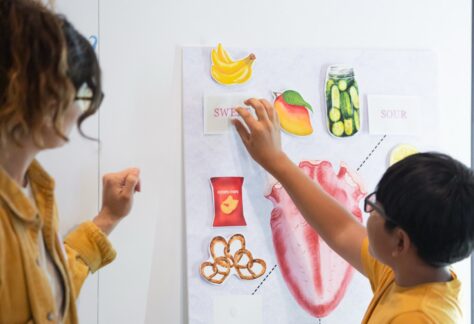 A young boy labels a tongue map with his teacher looking on