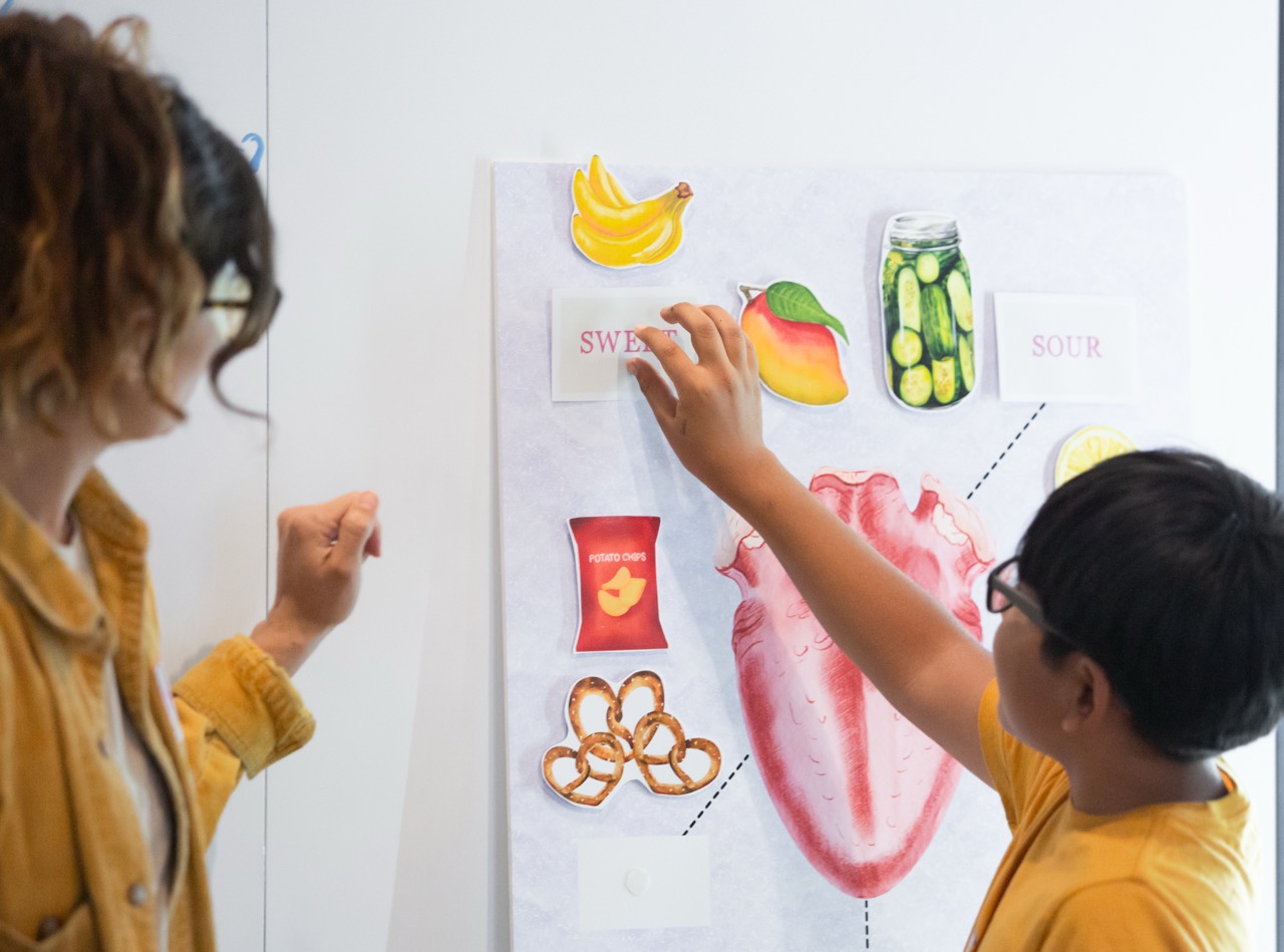 A young boy labels a tongue map with his teacher looking on